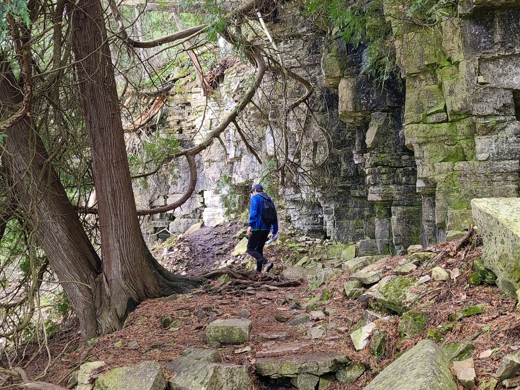 man hiking on a trail passing between trees and a rocky bluff