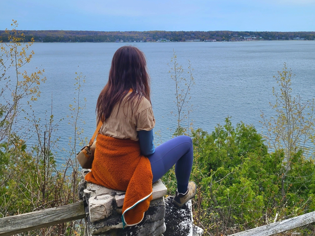 woman in hiking attire sitting on a rock pillar of a fence looking over a bluff onto a Lake Michigan shoreline